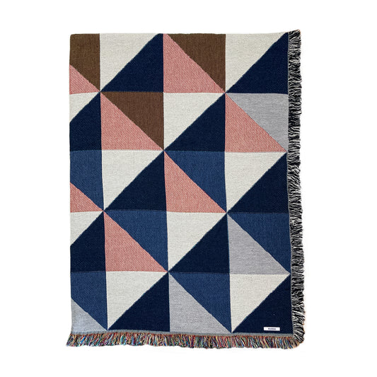 A woven cotton throw blanket with a geometric pattern in navy, pink, ochre, and gray, featuring a fringe edge.