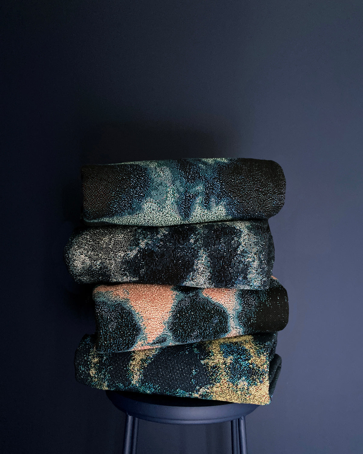Grouping of woven throw blankets, stacked on stool against navy background.